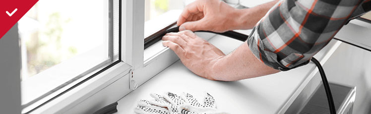  A person in a plaid shirt installs black weather stripping around the frame of a white window.
