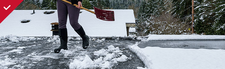 A person wearing dark pants and black boots stands on a flat, dark roof using a red shovel to clear away patches of white snow.