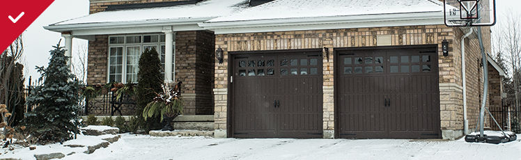 A snow-covered brick house features two dark brown garage doors, a snow-dusted driveway, and a basketball hoop to the right.