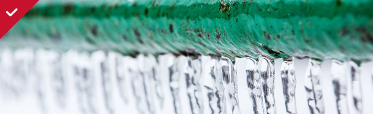 A close-up view shows many small, clear icicles hanging in a row from the bottom of a weathered green metal pipe.