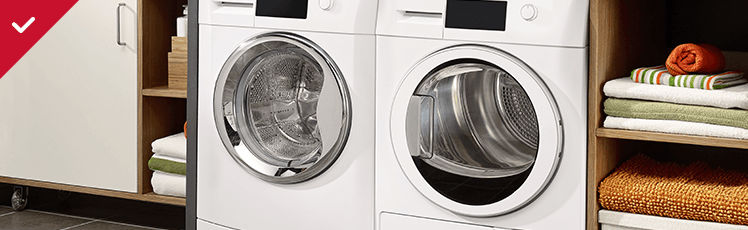  A white washing machine and matching dryer are positioned side-by-side next to open wooden shelves filled with neatly folded towels in shades of white, green, and orange