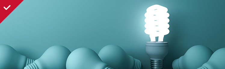  A glowing white spiral compact fluorescent light bulb stands upright amidst a group of matte teal traditional light bulbs lying on their sides.