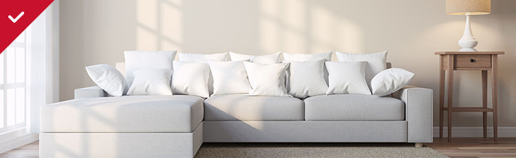 A modern light-gray sectional sofa topped with numerous white pillows sits against a beige wall in a brightly lit room next to a wooden side table with a white lamp.