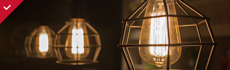 : Three industrial-style pendant lights with glowing Edison-style bulbs are enclosed in black wire cages against a dark background.