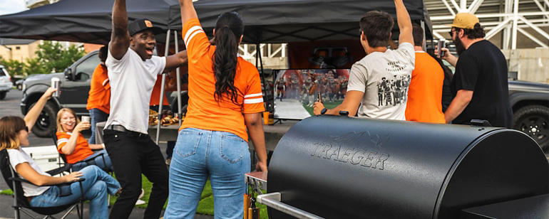 Tailgate party with multiple people wearing orange jerseys celebrating and a Traeger pellet grill in the foreground