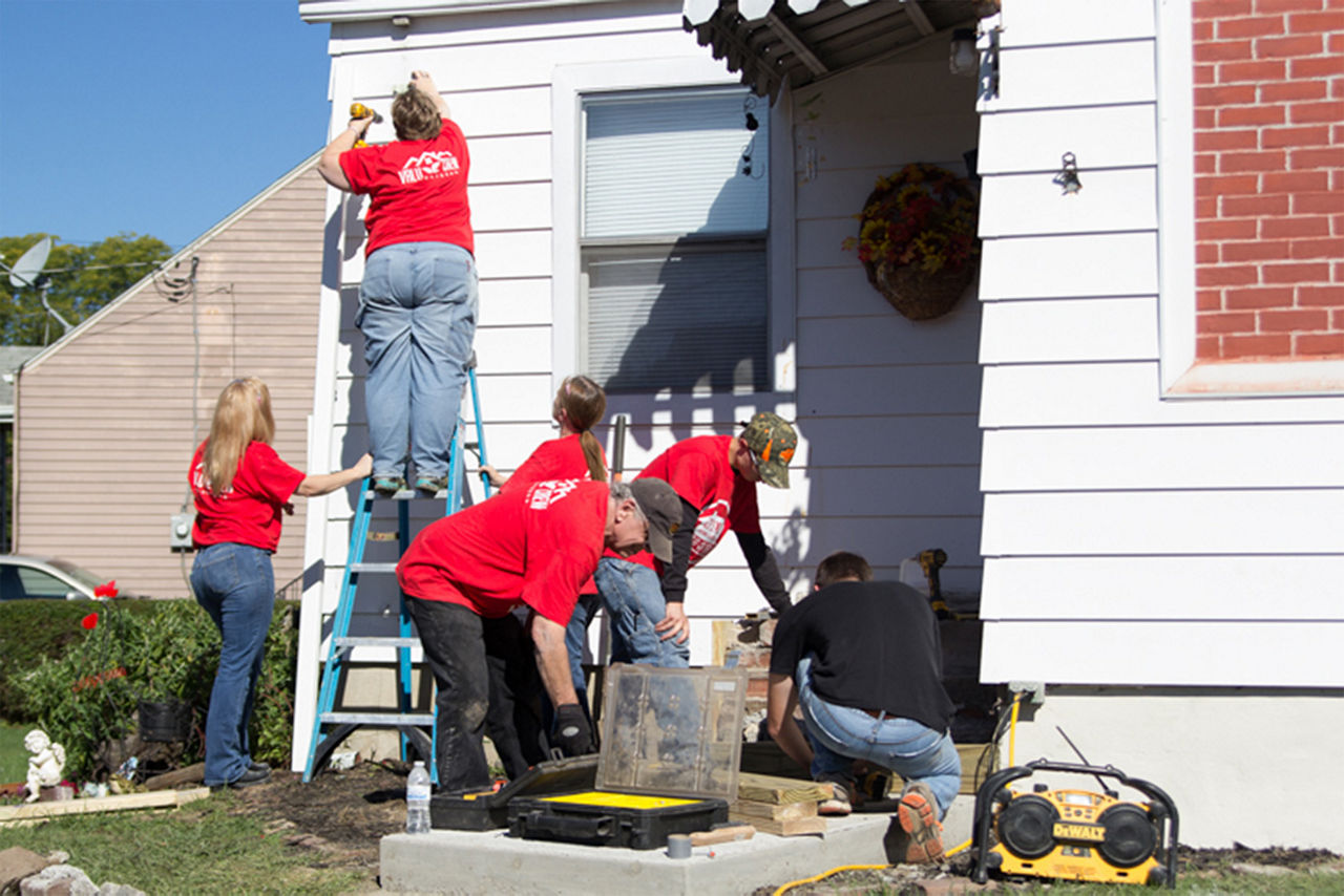 A woman on a ladder drilling into the house while people are around