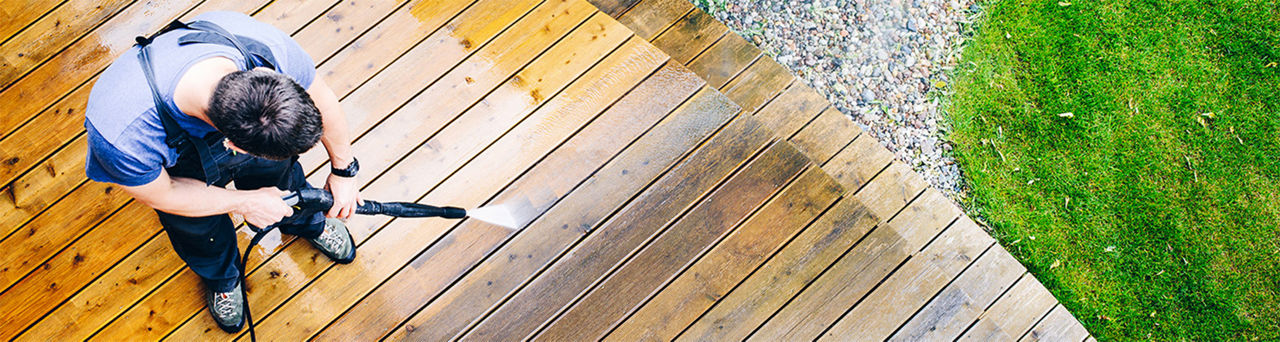 A top view of a man in a blue shirt and overalls pressure washing a dirty deck 