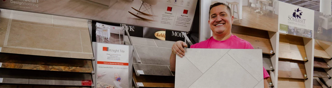 A man wearing a pink shirt is holding up a piece of tile flooring