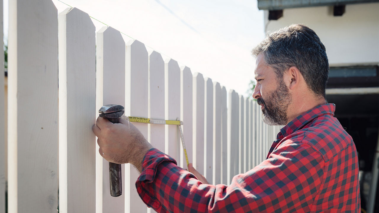 Carpenter craftsman with electric sander smoothes the boards of a wooden fence.