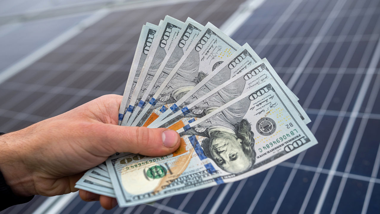 A hand holding money in front of solar panels 