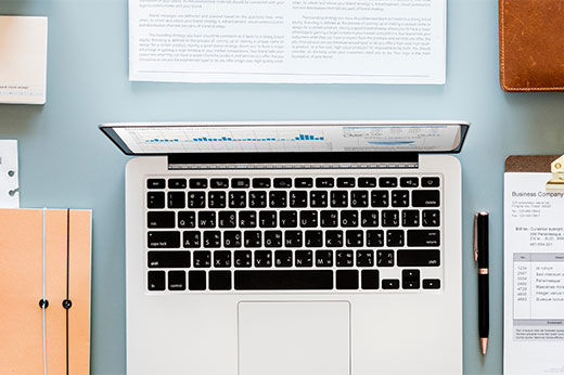 A top down view of an office desk with a laptop, clipboard, stack of papers, coffee cup, glasses, phone, and a brief case