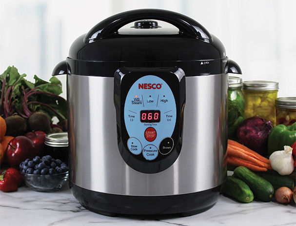 A front view of a water pressure cooker that is silver with a black top and bottom. There are a variety of vegetables on the table behind the cooker