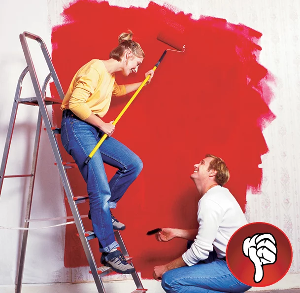 a couple painting an interior wall red. The women is leaning on a step ladder holding a paint roller and cover while the man is kneeling on the ground painting around the trim