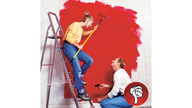 a couple painting an interior wall red. The women is leaning on a step ladder holding a paint roller and cover while the man is kneeling on the ground painting around the trim