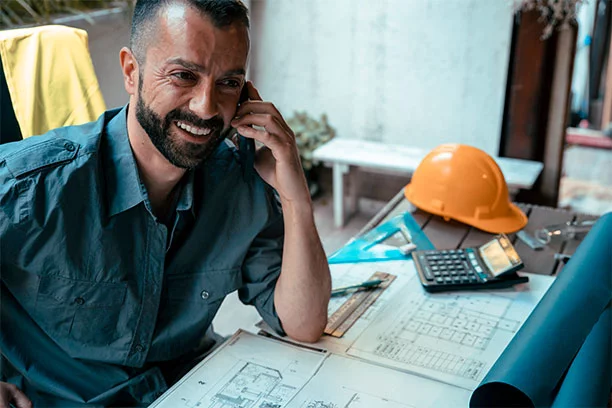 Man on the phone with blueprints, calculator, triangle, and hard hat on his desk