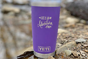 Purple Yeti Tumbler sitting outside on rocks with custom engraving that says "Life is an adventure" 
