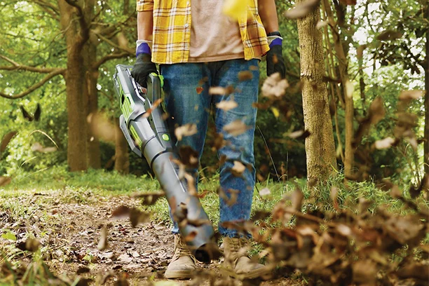 A man wearing a yellow shirt, jeans and black gloves, using a battery powered leaf blower to clear away fallen leaves 