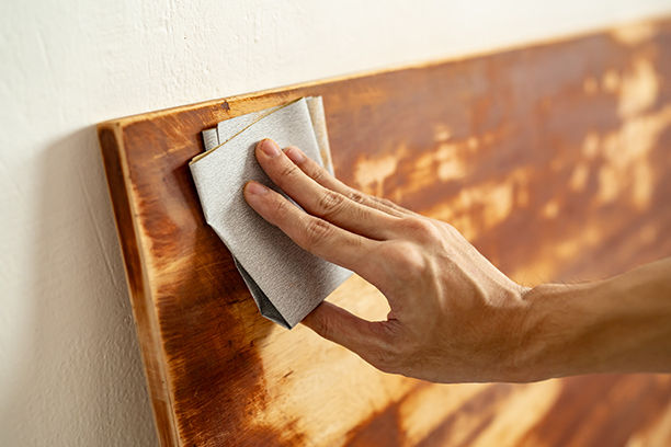 Person using sandpaper to sand a piece of wood against a neutral colored wall