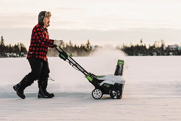 A man wearing a red plaid flannel, black snowpants, black snow boots, and a black fuzzy hat pushing a green and black snow blower in the snow