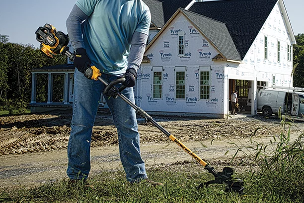 Man using a Dewalt battery powered string trimmer to clear away brush in front of a home construction site 