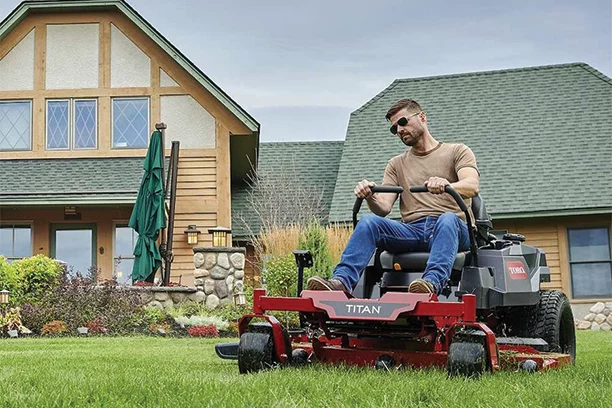 Middle aged man wearing a tan shirt and jeans using a red zero turn mower to mow his backyard. There is a beige and white house behind him with green shingle roofing 