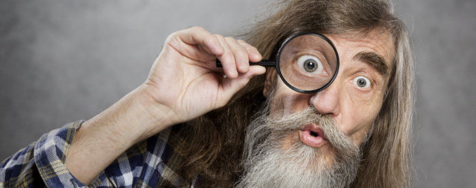 An old man with long hair and a beard holding a magnifying glass held up to this right eye