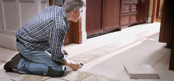 Man kneeling on the ground using a Bosch laser distance measurer to measure the distance of tiles in a bathroom