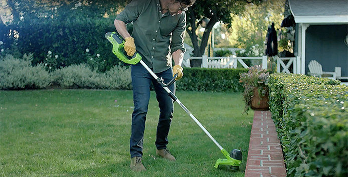 A person wearing a dark green button-down shirt, blue jeans, brown work boots, safety gloves, and goggles uses a lime-green garden edger alongside a brick path on the edge of a green lawn. 