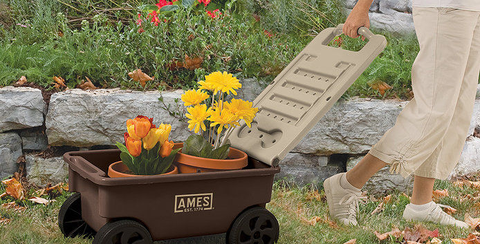 A person in khaki-colored pants and sneakers pulls a garden cart with one hand. In the black and khaki colored cart are two potted plants with orange and yellow flowers.