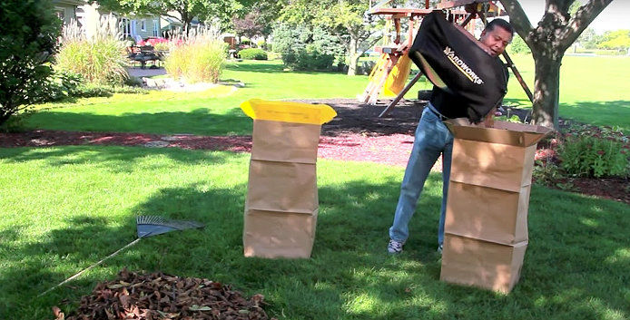 A man pours a black lawnmower bag into a brown, standing leaf collection bag. The man is working in a yard of green grass with trees and bushes in the background and a rake near his workspace. 