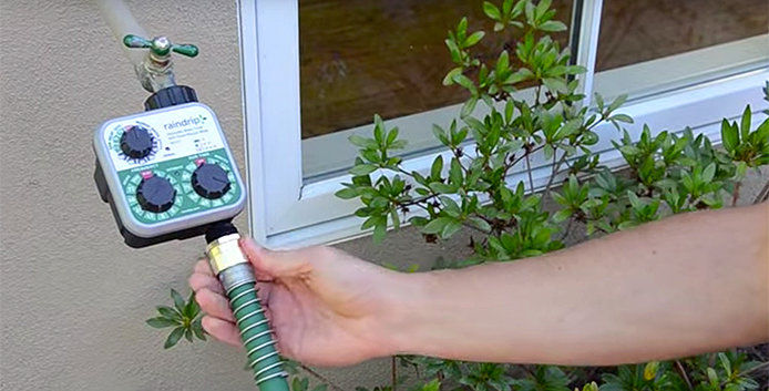 A person is attaching a green garden hose to a green hose timer box with black knobs. 