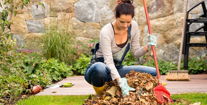 A brunette woman in a grey long-sleeved shirt and jeans is wearing mint-green garden gloves. The woman crouches down and uses an orange rake to scoop up brown leaves from the green grass.