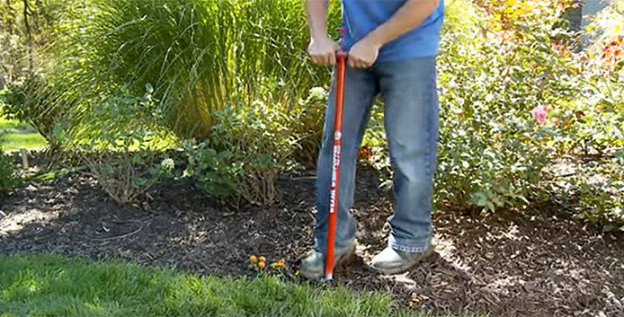 A person in a blue shirt and jeans uses a red-handled yard tool to edge a garden bed. 