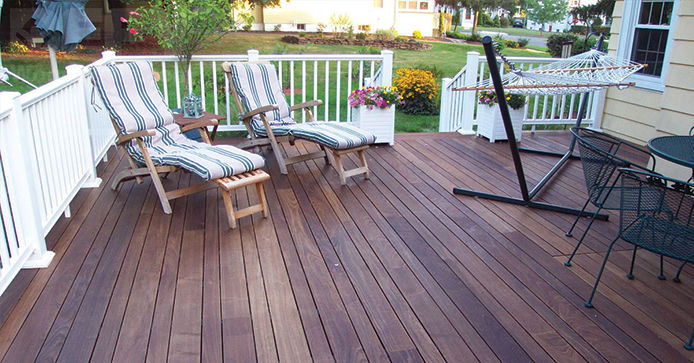 A dark-stained wooden deck is surrounded by a white guardrail. The deck features two striped lounge chairs, a hammock, and several potted plants. 