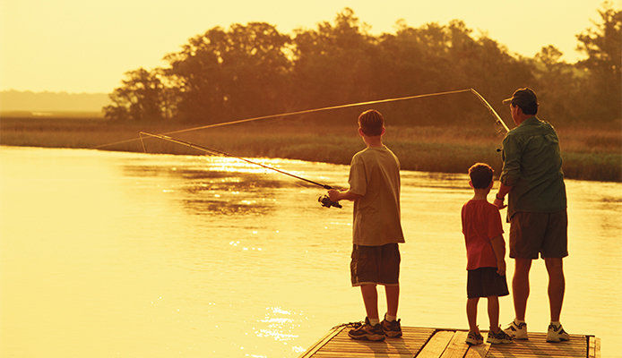 An adult and two younger people stand on a dock, casting fishing lines into a lake at sunset. The fishermen are wearing shorts and dressed for warm weather, and the lake appears calm with a golden yellow glow. 