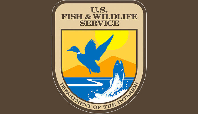 The US Fish & Wildlife Service insignia on a brown background. The logo depicts a blue duck against an orange skyline, with a trout jumping out of the water below and reads "Department of the Interior" across the bottom. 
