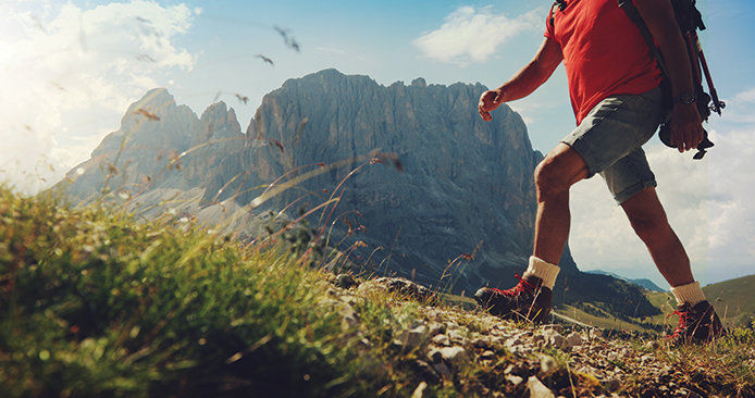 A man wearing a red shirt and gray hiking short, while carring a backpack is hiking up a grassy hillside. There is a rock formation in the back ground and the sky is blue.