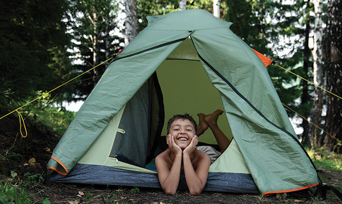 A young smiling boy is laying half way in and half way outside of a green camping tent. 