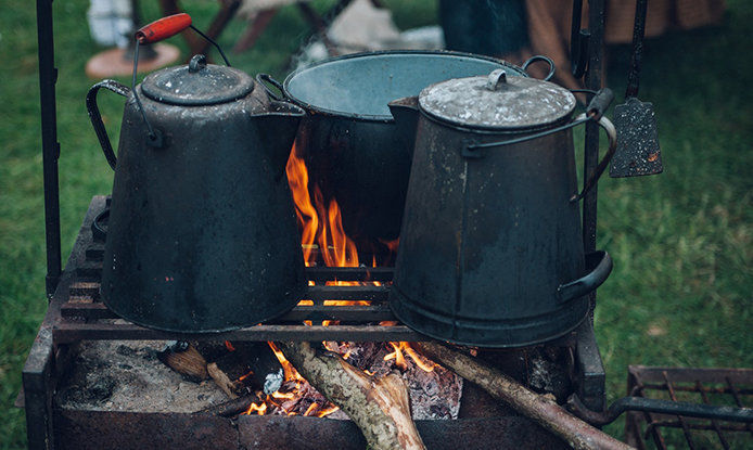 3 cast iron pots and pans sitting on a grate over a campfire. The fire is a blaze and there is wood sticking out of the firepit below the grate.