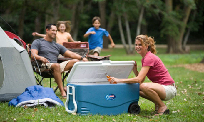 A family at a campground smiling having a good time. The mother is squatting down in front of a 40 gallon blue cooler. She is pulling out a orange soda in a glass bottle while the father and kids are in the background hanging out in camping chairs.