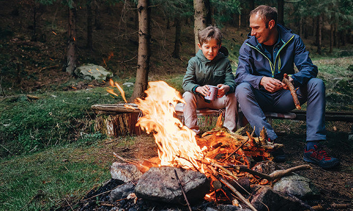 Fatehr and son sitting in front of a blazing camp fire holding a cup of hot chocolate camping in the woods. 