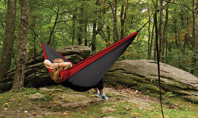 a woman relaxing inside of a red and gray hammock. The hammock is hung in the woods between two trees. There is a rock formation in from of the hammock.