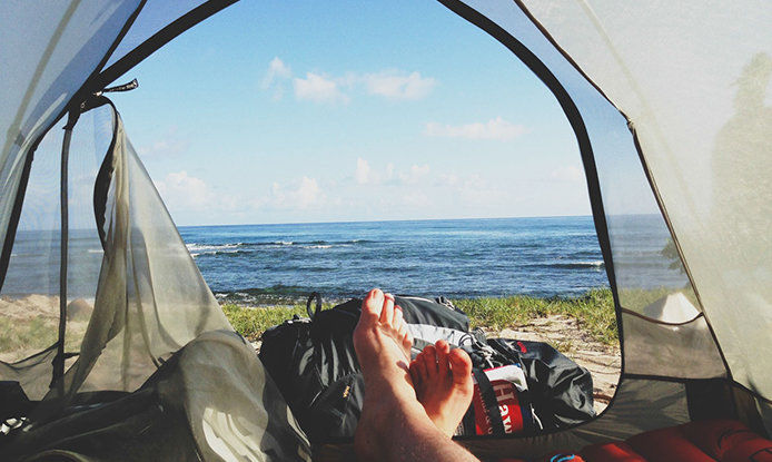 A man laying inside of a tent that is facing the ocean. The tent is slightly open and you can only see the toes of the man laying down. There is also a camping bag at the opening of the tent. 