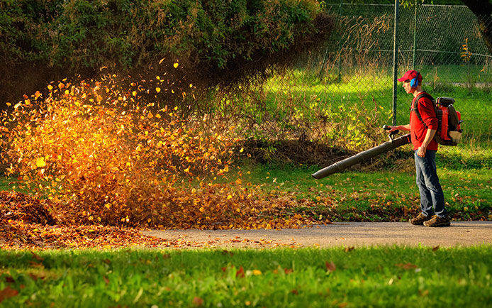 Man in red wearing ear protection using a leaf blower to blow leaves off of the sidewalk