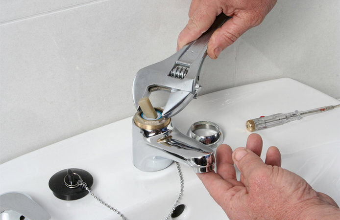 A person repairing a faucet using a silver wrench to tighten the top of the faucet. The person is repairing a silver-colored faucet on a white ceramic sink.