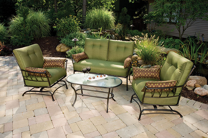 five piece patio set with green cushions and a glass table top sitting on a stone patio 