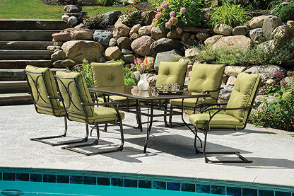 A seven piece patio furniture set with a glass top table and green cushions sitting on a stone patio next to a pool