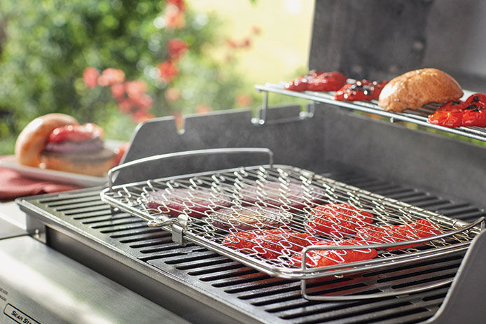 A close up of a grill basket holding sliced onions and tomatoes set on top of a gas stove