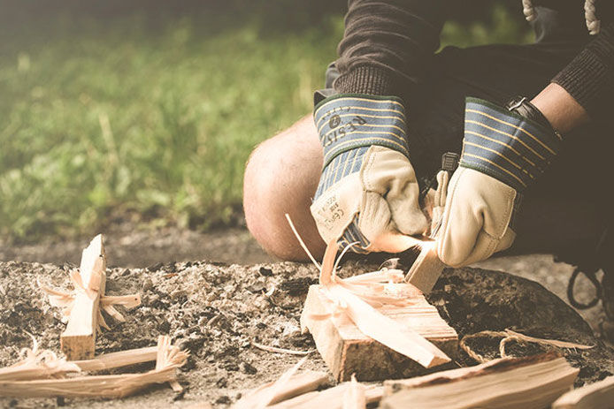 A man making kindling from a log using a knife while wearing protective gloves