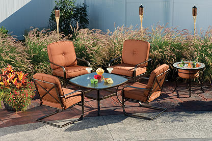 A six piece patio set with glass table top sitting on a stone patio surrounded with ornamental grass and tiki torches